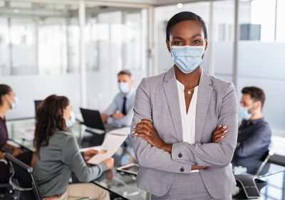 Portrait of african american businesswoman wearing protective face mask standing in modern office. Successful black entrepreneur in conference room leaning over table wearing surgical mask with business people working in background. Successful leader looking at camera with crossed arms at work.